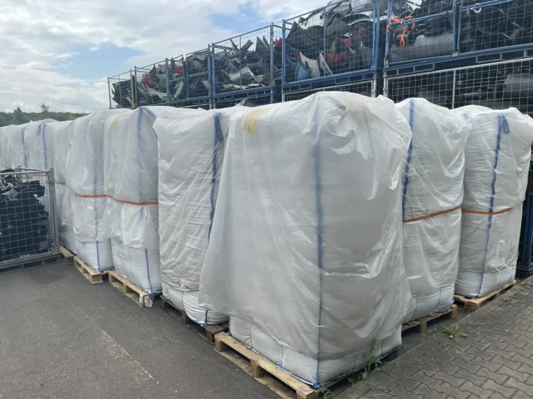 Reihe weißer Big-Bags mit Mahlgut auf Paletten, im Hintergrund Gitterboxen mit Fahrzeugteilen