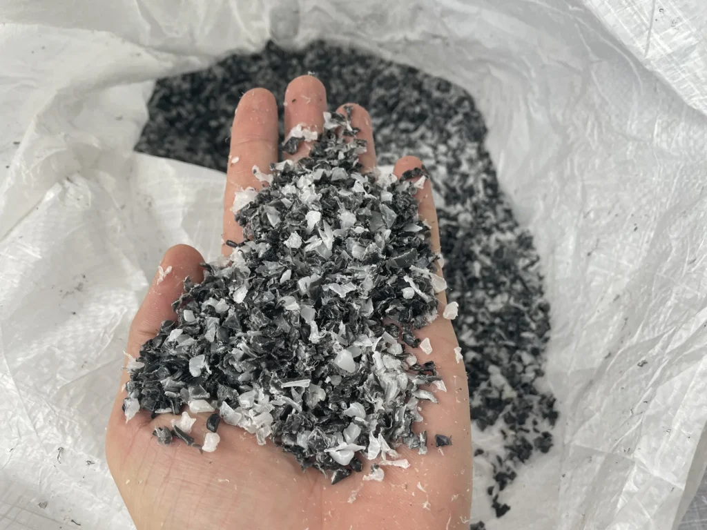 Dark grey plastic regrind with lighter flakes on an open hand above a big bag